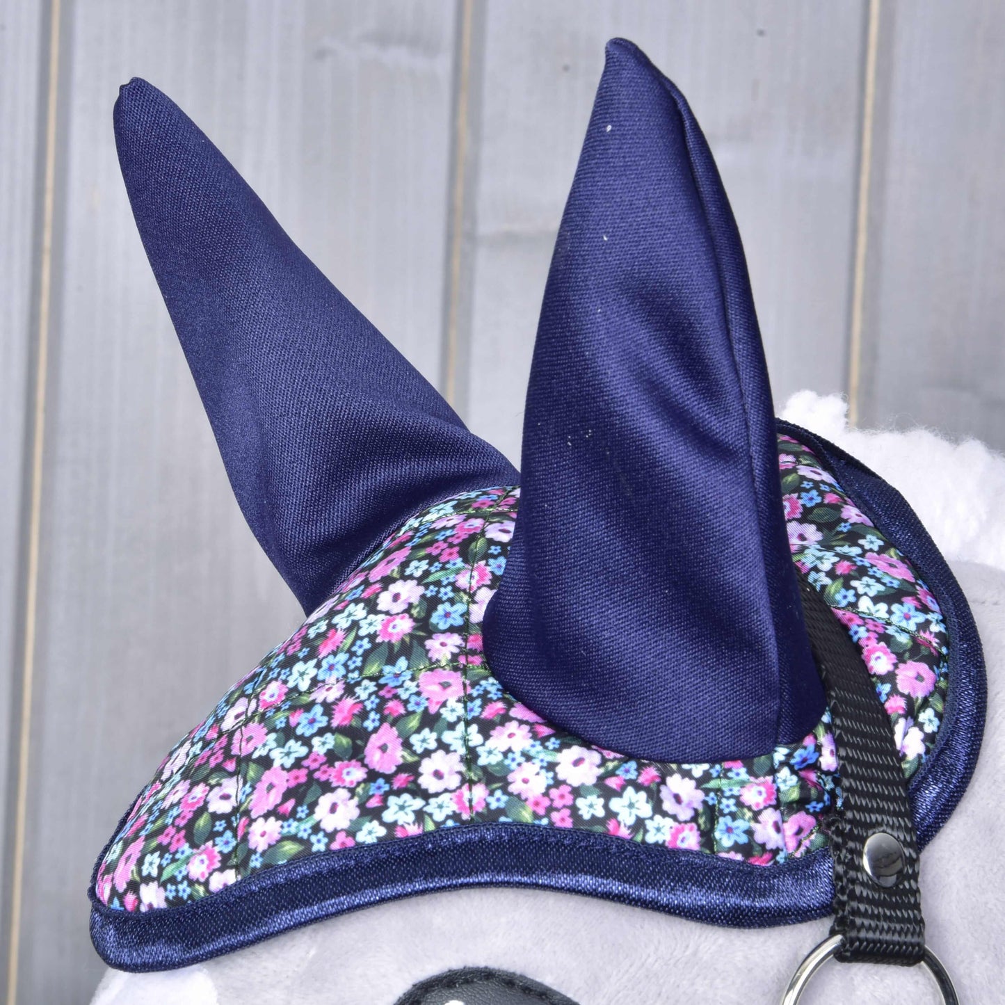 Gros plan sur un cheval portant le Bonnet d'oreilles Fleur de Hobby Horse LarDen, avec oreilles bleu foncé et motif floral rose, blanc et bleu ; une partie de la bride et le fond gris clair montrent sa protection élégante contre les insectes.