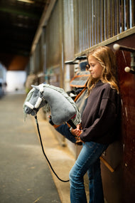 Mädchen in dunkelbraunem Sweatshirt lehnt in einer Stallgasse an den Boxen und hält das Hobby Horse Calypso mit schwarzem Halfter von Hobby Horse LarDen – grauer Plüschkopf, hellgraue Garnmähne – mit gefüttertem Teddy Classic Halfter und schwarzem Führstrick