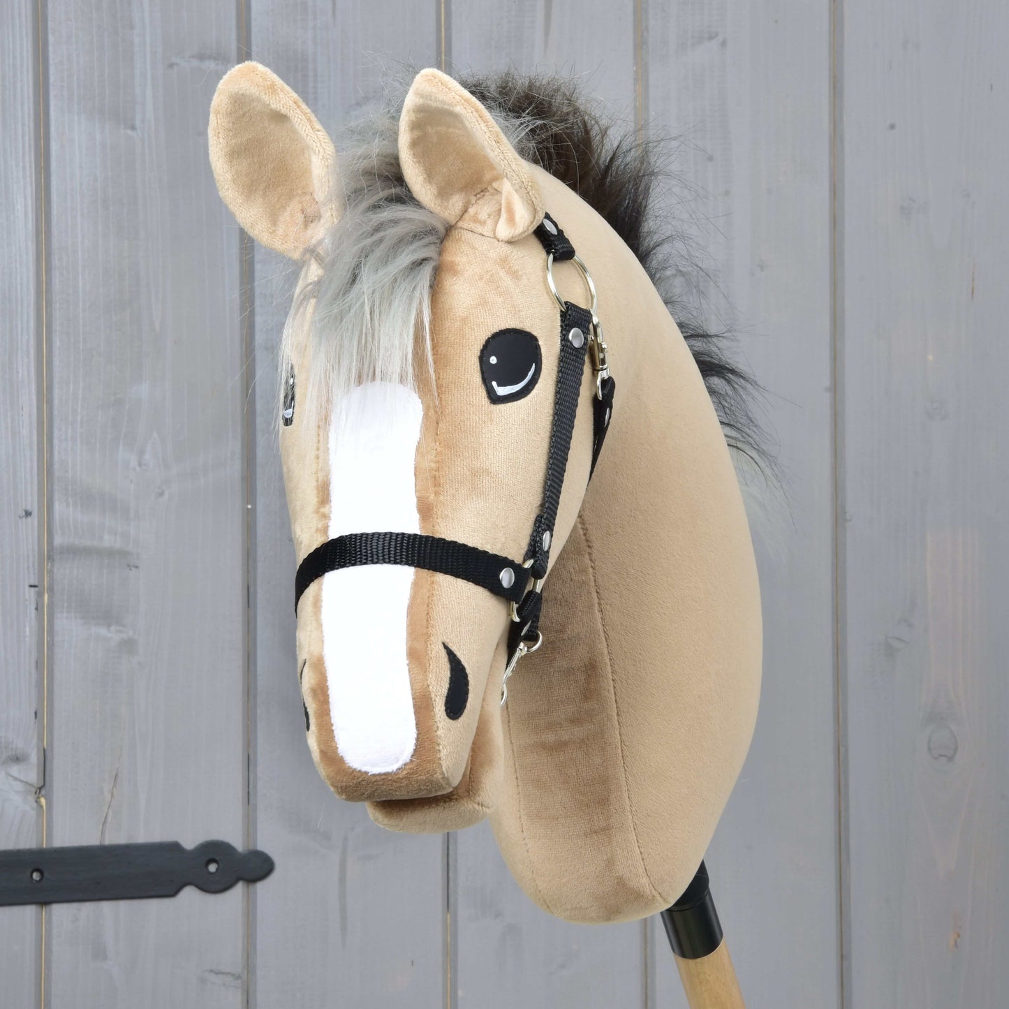 Hobby Horse Caramel avec licol noir de Hobby Horse LarDen, beige avec une liste blanche sur le visage et monté sur un bâton en bois, devant un mur en bois gris, dans le style d’un cheval de course Warmblood anglais.
