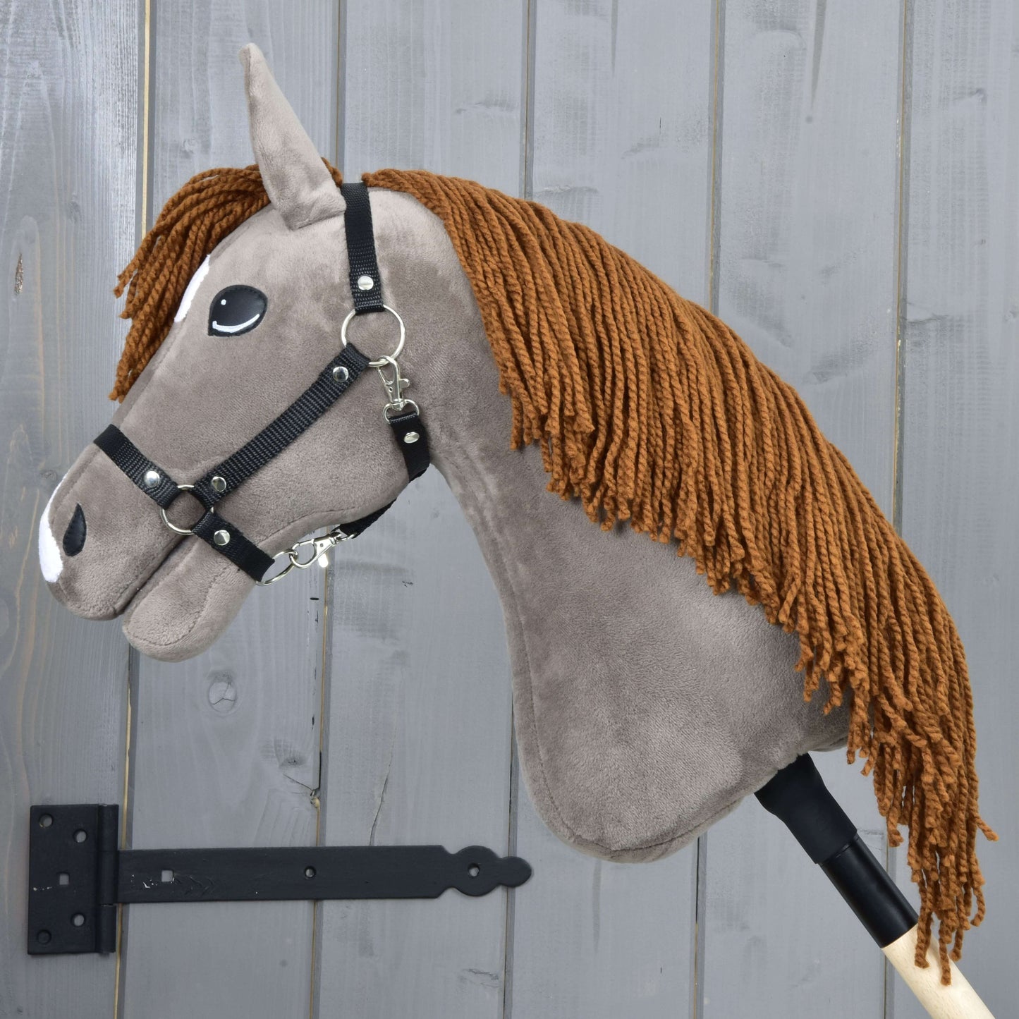 Hobby Horse Chanell avec licol noir de Hobby Horse LarDen, avec tête grise en tissu, crinière en fil brun, licol noir et accents blancs en feutre, monté sur un bâton en bois devant un mur en planches de bois grises.