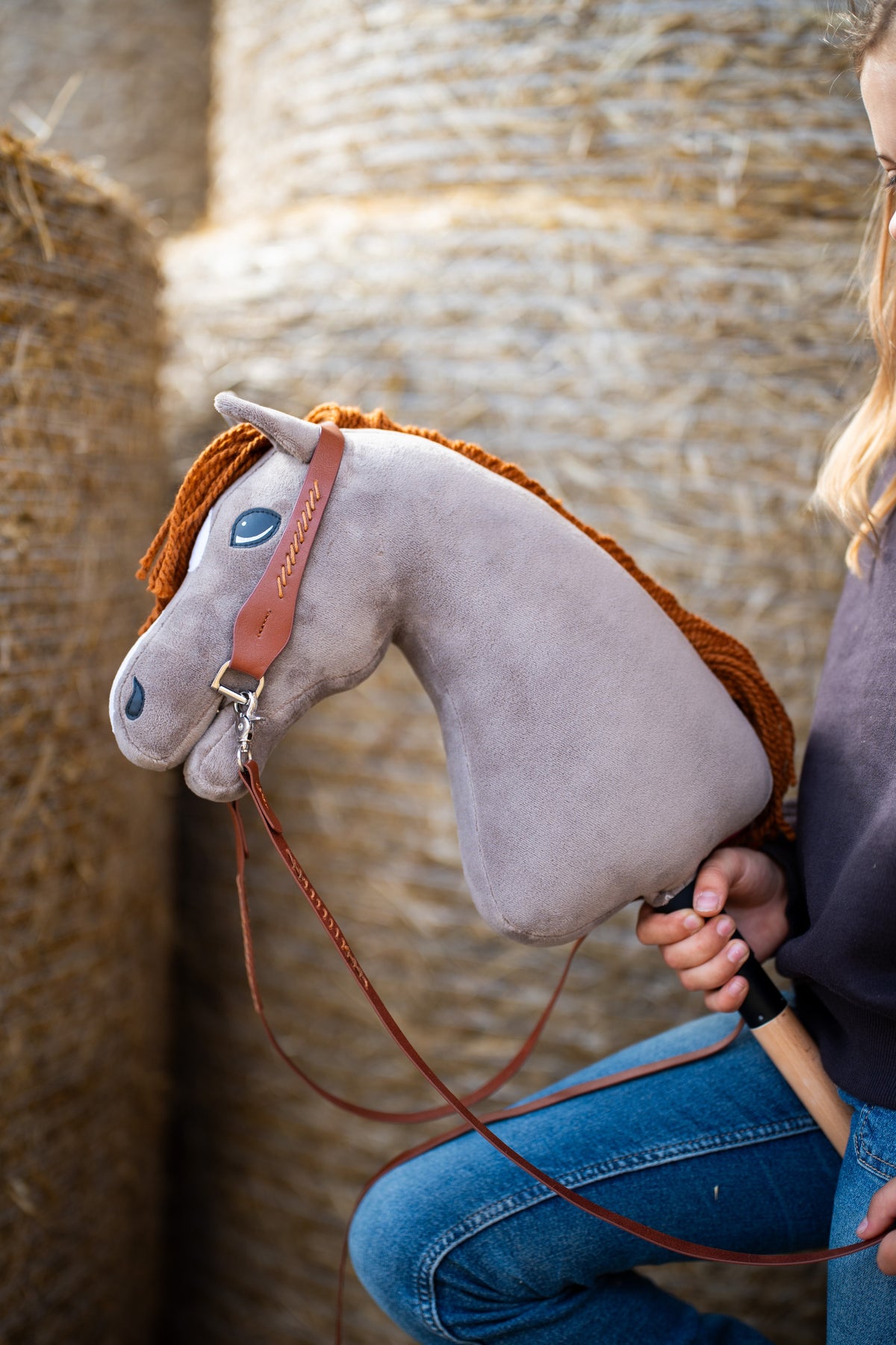 Kopf des Hobby Horse Chanell mit schwarzem Halfter von Hobby Horse LarDen vor runden Strohballen – grauer Plüsch, blaues Auge, ingwerfarbene Garnmähne – hier mit dem vollständigen Western-Zaumzeug mit einem Ohrteil aus braunem Leder