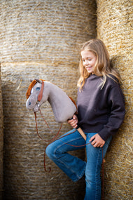 Lächelndes Mädchen im dunkelbraunen Sweatshirt lehnt an runden Strohballen und hält das Hobby Horse Chanell mit schwarzem Halfter von Hobby Horse LarDen – grauer Plüschkopf, blaues Auge, ingwerfarbene Garnmähne – hier mit dem vollständigen Western-Zaumzeug mit einem Ohrteil