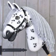 Hobby Horse LarDen "Hobby Horse Chantal mit Schwarzem Halfter" mit handgefertigtem weißem Kopf im Knabstrupper-Stil, schwarzen Flecken, grauer Garnmähne, schwarzem Halfter und Holzstabgriff vor einem Hintergrund aus hellen Holzplanken.