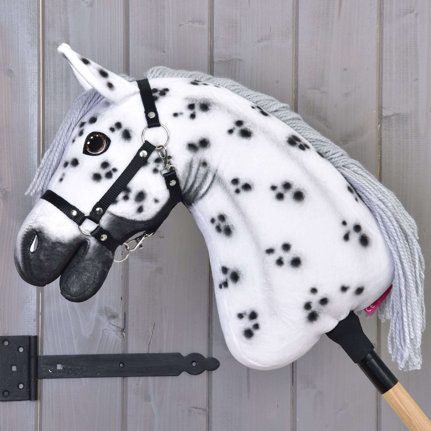 Hobby Horse Chantal avec licol noir de Hobby Horse LarDen, avec robe blanche à pois noirs, crinière grise en fil, licol noir et monté sur un bâton en bois devant un mur en bois gris clair.