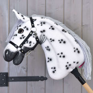 Hobby Horse LarDen "Hobby Horse Chantal mit Schwarzem Halfter" mit weißem Fell mit schwarzen Flecken, grauer Garnmähne und schwarzem Halfter, auf einem Holzstab vor einer hellgrauen Holzwand.