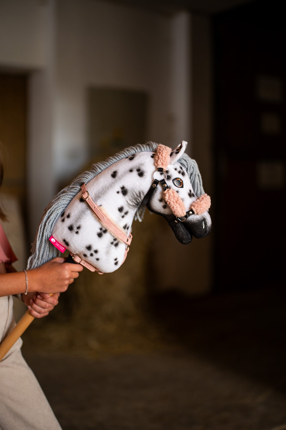 Kopf und Hals des Hobby Horse Chantal mit schwarzem Halfter von Hobby Horse LarDen werden im Stall hochgehalten – weißer Plüsch mit schwarzen Leopardenflecken, graue Garnmähne, schwarze Maulpartie – hier mit dem Old Rose Fellhalfter aus Kunstleder und dem Brustgeschirr mit Fell