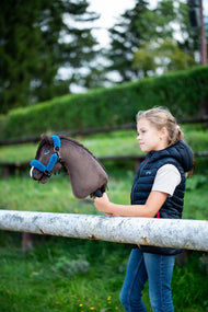 Lifestyle photo: a girl in a navy padded gilet stands behind a white paddock rail holding the Hobby Horse Chianti with Black Halter by Hobby Horse LarDen, a clipped green hedge behind - dark brown plush head, dark yarn mane - shown here with the fur-lined Teddy Petrol halter.