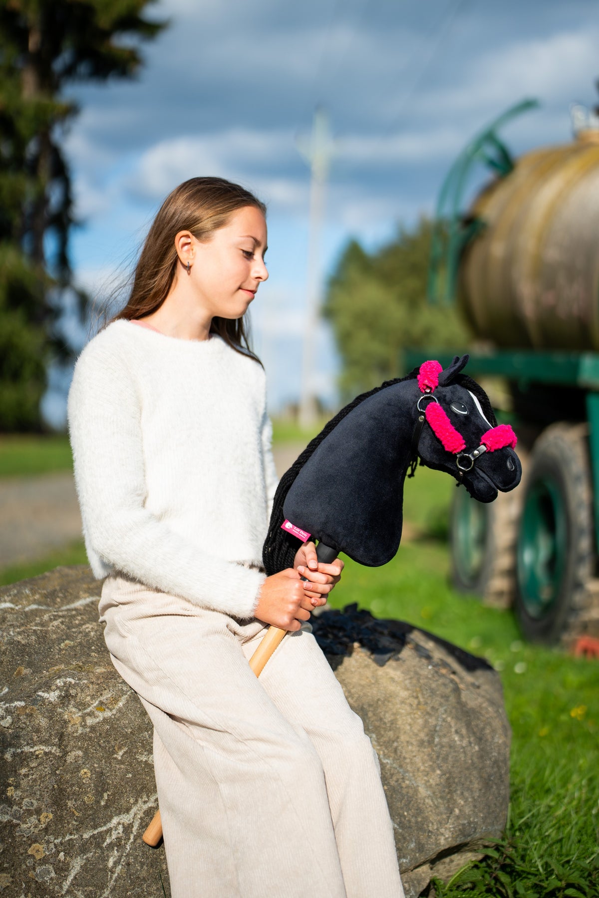 Mädchen im cremefarbenen Pullover sitzt auf einem Felsblock an einem Wiesenweg und hält das Hobby Horse Cloony mit schwarzem Halfter von Hobby Horse LarDen – schwarzer Plüschkopf, weißer Stern, schwarze Garnmähne – hier mit dem schwarzen Kunstlederhalfter mit vollständigem rosa Fell
