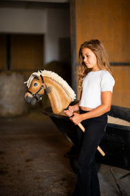 Mädchen im weißen Oberteil steht im Stall und hält das Hobby Horse Colby mit schwarzem Halfter von Hobby Horse LarDen – Palomino-Plüschkopf, dichte cremefarbene Garnmähne, schwarzes Halfter mit Silberbeschlägen