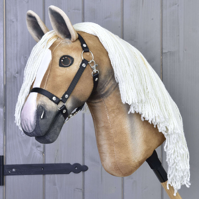Hobby Horse Connie mit Schwarzem Halfter von Hobby Horse LarDen mit realistischem hellbraun-weißem Design und langer weißer Garnmähne, auf einem Stock befestigt vor einer grauen Holzwand.