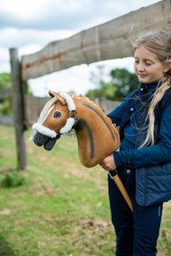 Mädchen in dunkelblauer Weste steht an einem hölzernen Koppelzaun und hält das Hobby Horse Connie mit schwarzem Halfter von Hobby Horse LarDen – Fuchs-Plüschkopf mit dunklem Aalstrich, cremefarbene Garnmähne – hier mit dem fellgefütterten Teddy Classic Halfter