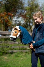 Mädchen in dunkelblauer Weste hält das Hobby Horse Connie mit schwarzem Halfter von Hobby Horse LarDen in einer petrolblauen Stalldecke, dahinter Herbstbeeren – Fuchs-Plüschkopf, cremefarbene Garnmähne – hier mit dem fellgefütterten Teddy Classic Halfter und der Winter Sky Stalldecke