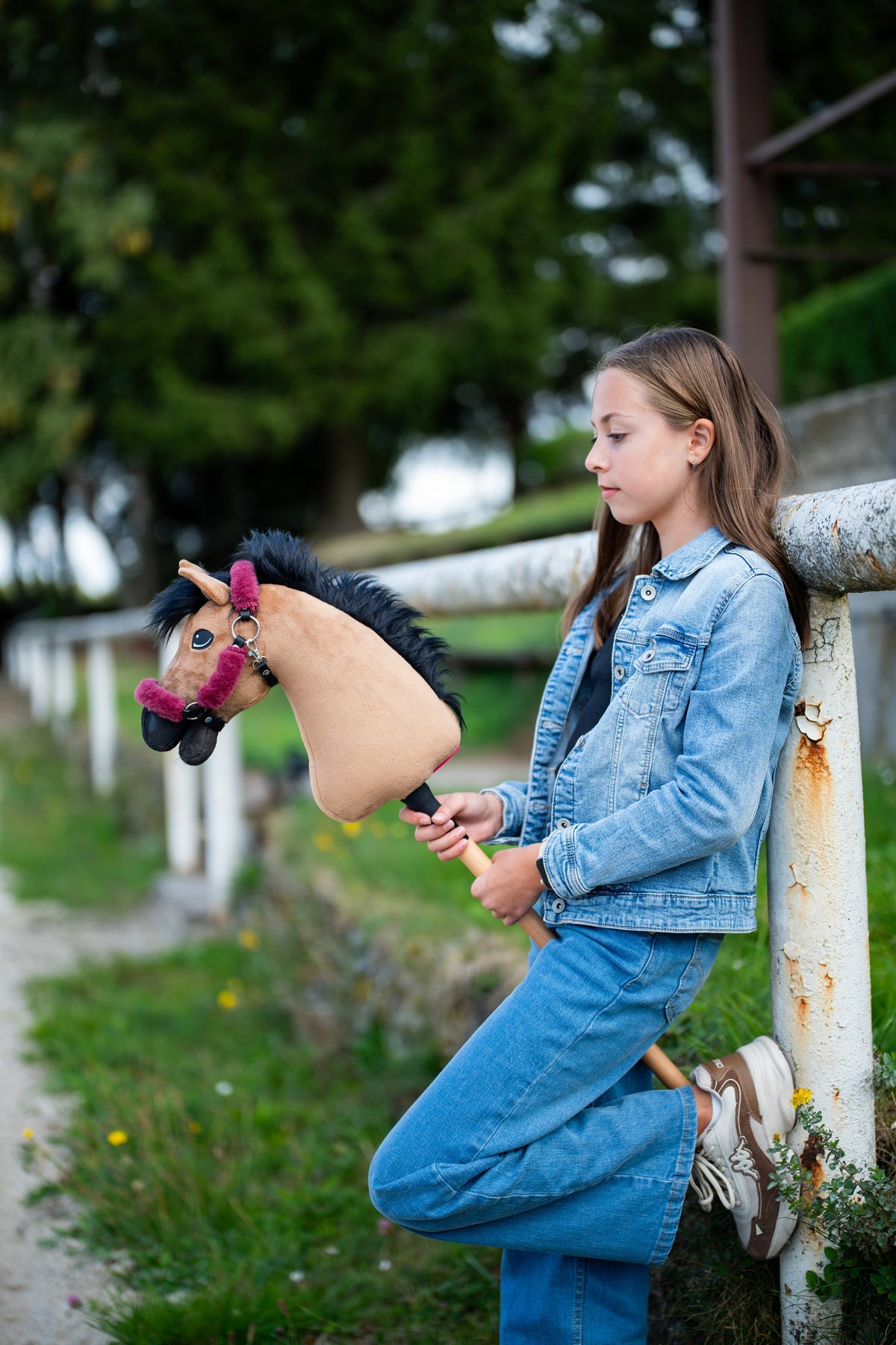 Mädchen im Jeanslook lehnt an einem weißen Koppelzaun und hält das Hobby Horse Cookie mit schwarzem Halfter von Hobby Horse LarDen – sandfarbener Plüschkopf, schwarze Garnmähne – hier mit dem fellgefütterten Teddy Maroon Halfter