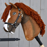 Hobby Horse Cooper mit schwarzem Halfter von Hobby Horse LarDen, ein braunes Hobby Horse inspiriert von einem Oldenburger Sportpferd. Dieses charmante Spielzeug hat eine weiße Blesse im Gesicht, dichtes dunkelbraunes Garnhaar und ein schwarzes Nylonhalfter mit Metallbeschlägen. An einem Holzstab befestigt und vor grauen Holzplanken abgebildet, ideal als fantasievolles Dressurspiel.