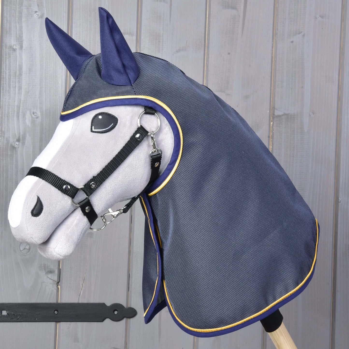 Die Hobby Horse LarDen „Regendecke Crone“ zeigt einen grauen Hobby Horse Kopf aus Plüsch mit schwarzem Zaumzeug und einer dunkelblauen Regendecke mit gelber Einfassung vor Holz-Hintergrund.
