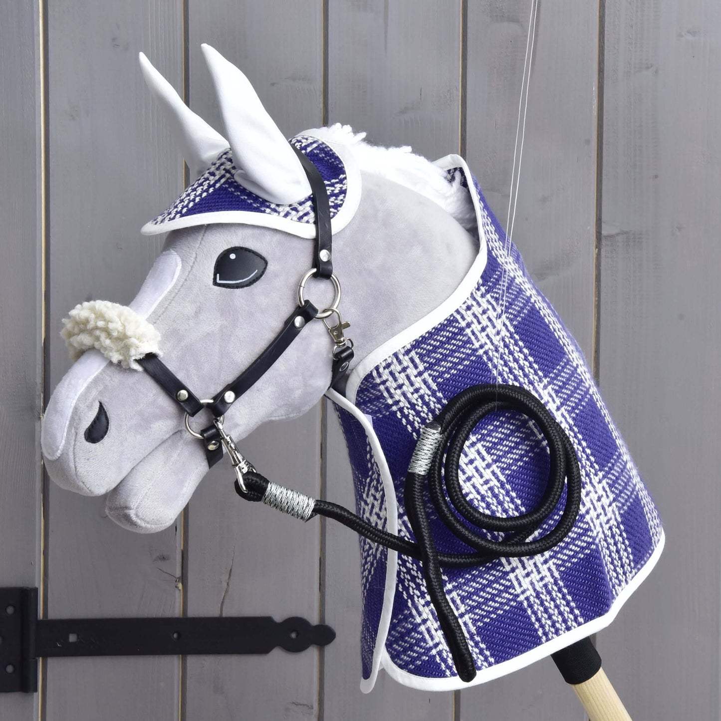 Ensemble Cube (couverture, bonnet d'oreilles, longe, licol avec fourrure) de Hobby Horse LarDen, comprenant une couverture, un bonnet d’oreilles, une longe et un licol en fourrure, présenté sur un mur en bois près d’une charnière de portail noire. Les accessoires mettent en valeur la qualité et le charme des produits Hobby Horse LarDen.