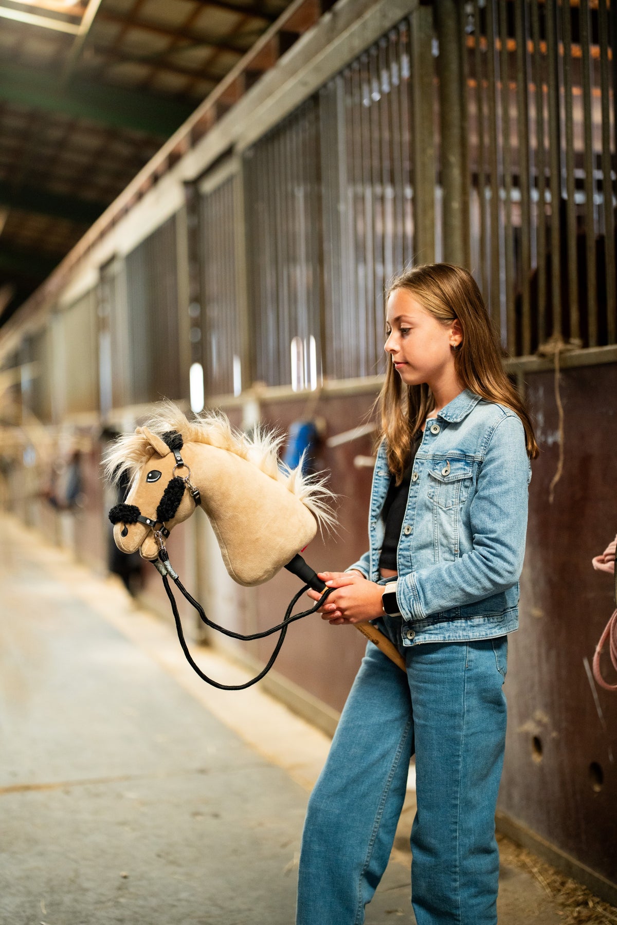 Mädchen in Jeansjacke führt das Hobby Horse Dancing Lady mit schwarzem Halfter von Hobby Horse LarDen durch einen Stallgang – cremefarbener Plüschkopf, helle Garnmähne und Stirnschopf – hier mit dem schwarzen Kunstlederhalfter mit vollständigem schwarzem Fell und schwarzen Zügeln