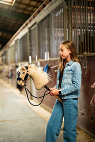 Mädchen in Jeansjacke führt das Hobby Horse Dancing Lady mit schwarzem Halfter von Hobby Horse LarDen durch einen Stallgang – cremefarbener Plüschkopf, helle Garnmähne und Stirnschopf – hier mit dem schwarzen Kunstlederhalfter mit vollständigem schwarzem Fell und schwarzen Zügeln