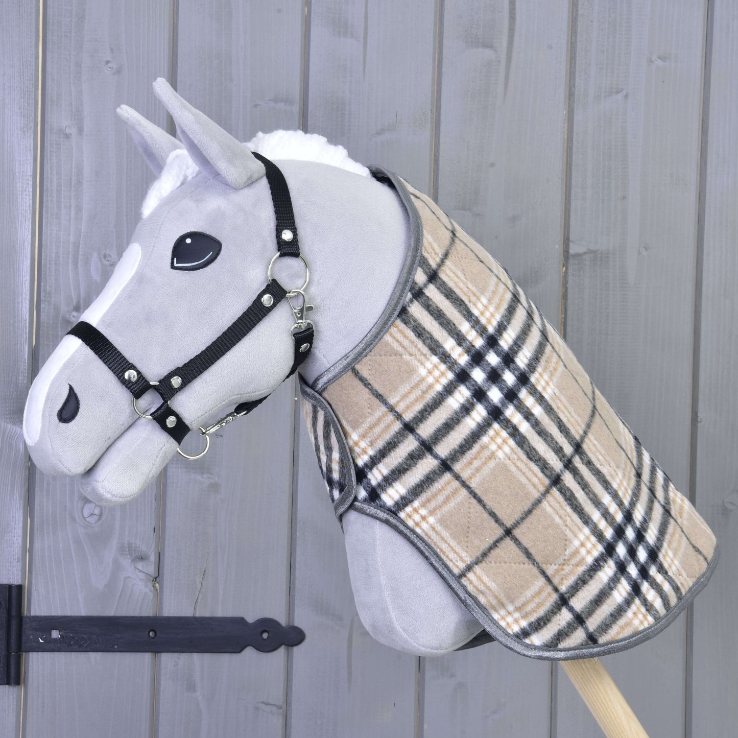 Die Hobby Horse LarDen "Decke Cremo England" ist ein graues Hobby Horse mit schwarzem Halfter für Hobby Horse und beigefarbenem kariertem Mantel, mit einer Fliegenhaube zum Insektenschutz, auf einem Holzstab vor einer grauen Holzwand.
