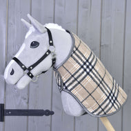Die Hobby Horse LarDen "Decke Cremo England" ist ein graues Hobby Horse mit schwarzem Halfter für Hobby Horse und beigefarbenem kariertem Mantel, mit einer Fliegenhaube zum Insektenschutz, auf einem Holzstab vor einer grauen Holzwand.