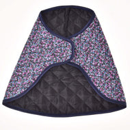 The Blanket Flower by Hobby Horse LarDen is a quilted, cape-style garment with black lining, a dark floral pattern of small blue and purple flowers, and dark blue trim—evoking a cozy horse blanket.