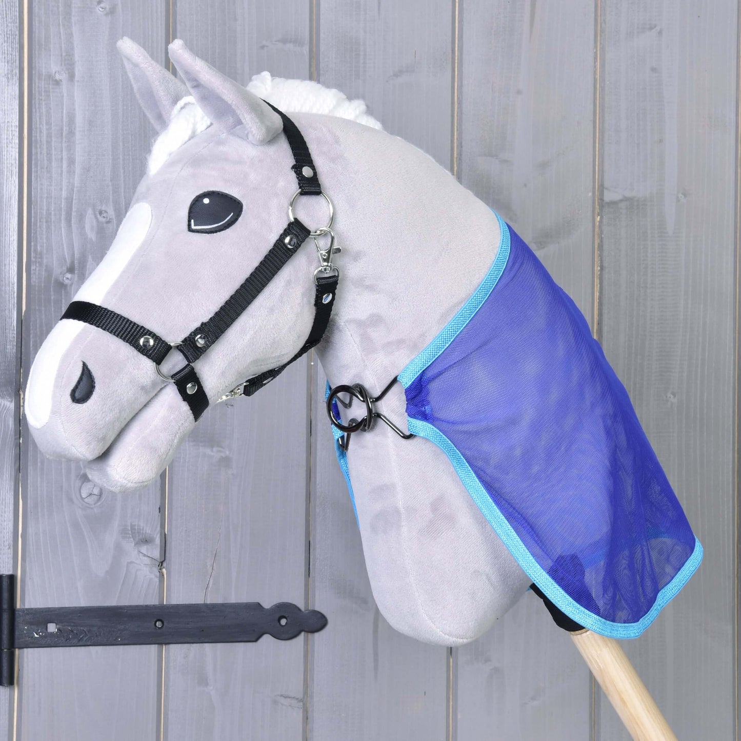 Ein graues Spielzeug von Hobby Horse LarDen mit schwarzem Zaumzeug und weißer Mähne trägt eine blaue Fliegendecke als Umhang. Das Pferd hat comicartige Augen und ist vor einer Holzwand montiert.