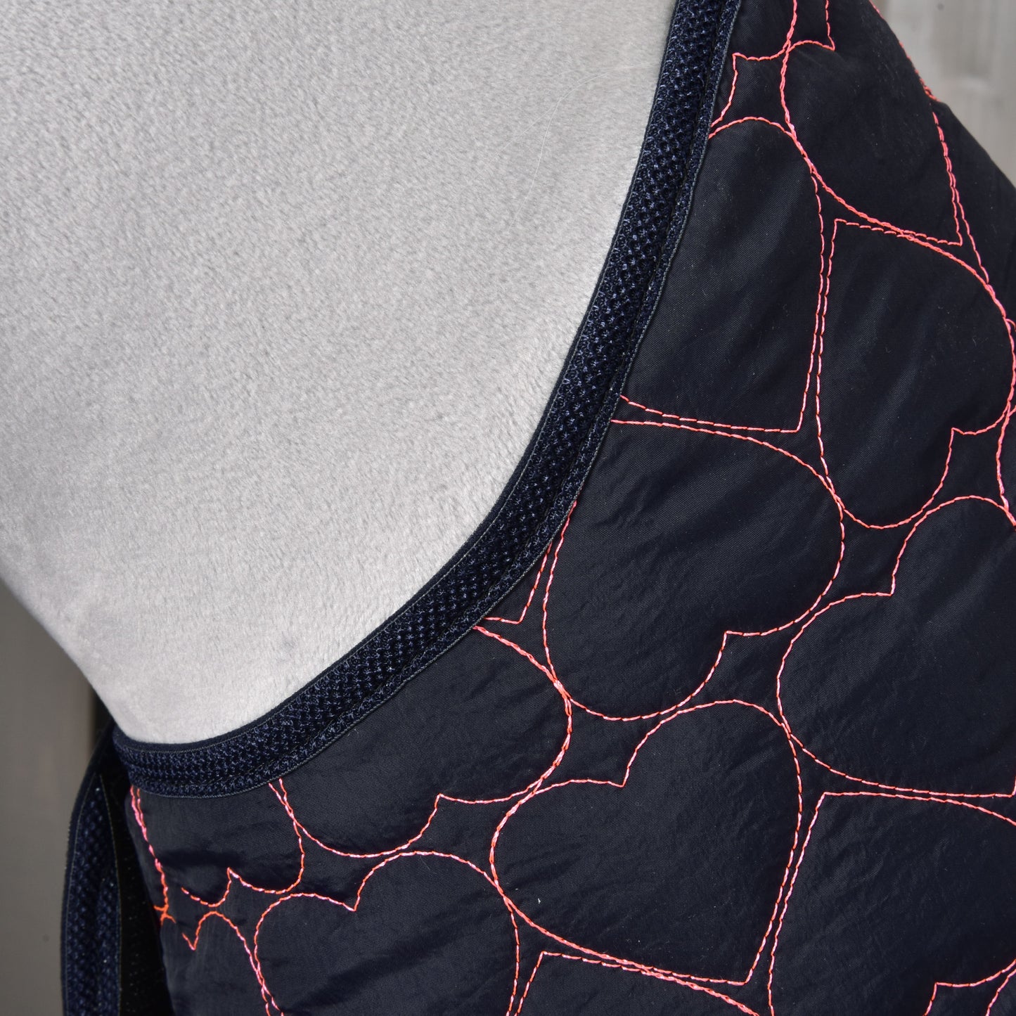Close-up of the Hobby Horse LarDen “Blanket Pink Heart,” a dark fabric horse training blanket featuring pink heart-shaped stitching patterns and dark blue trim, draped over a light grey surface.