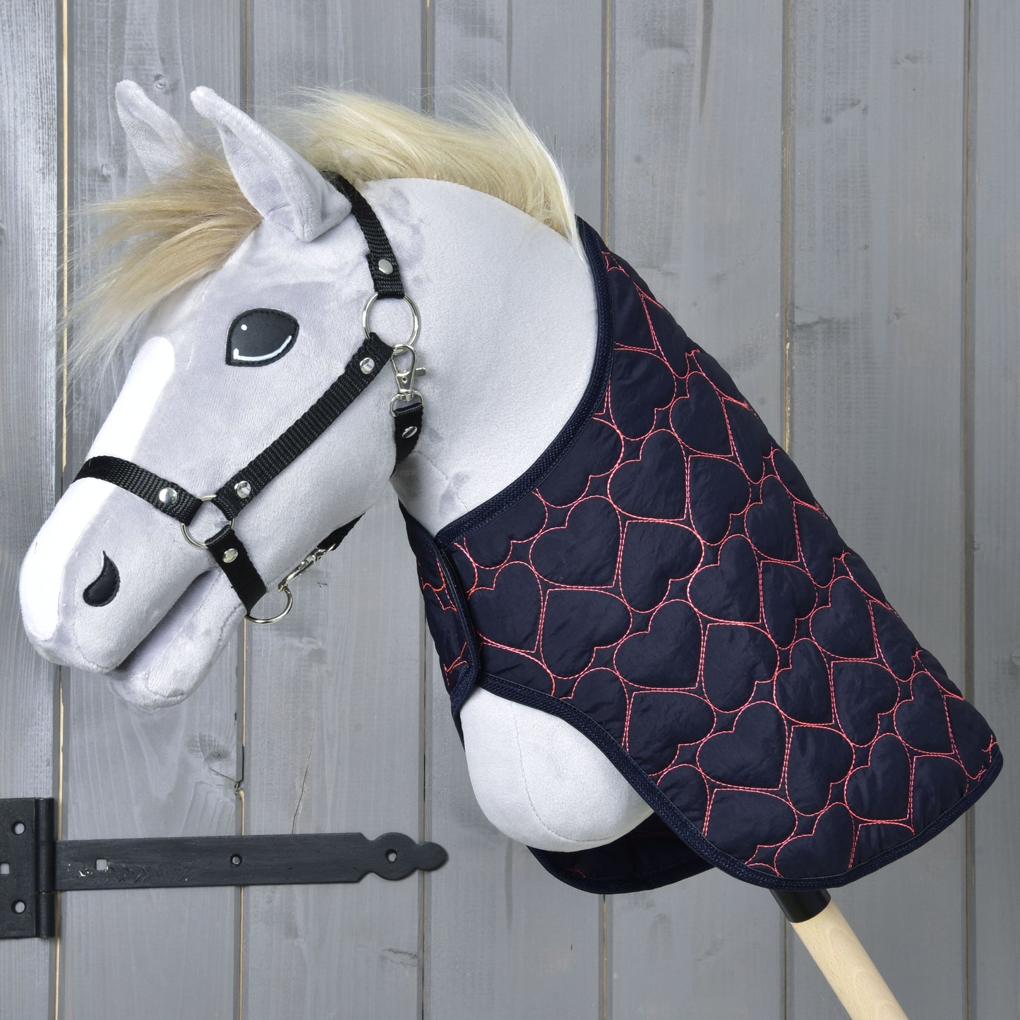 A plush hobby horse with a black halter and the Hobby Horse LarDen “Blanket Pink Heart” quilted hobby horse blanket, decorated with pink hearts, is posed against a wooden background.