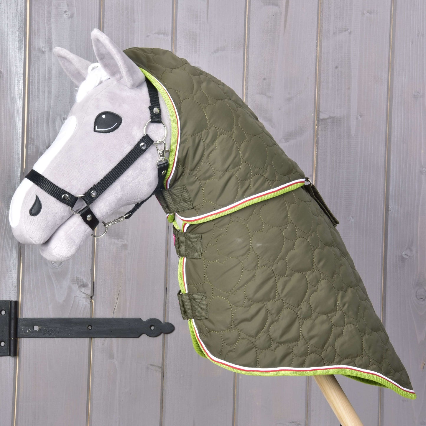 Ein graues Hobby Horse mit schwarzem Zaumzeug trägt die Decke mit abnehmbarem Halsteil Rubby von Hobby Horse LarDen, eine grüne, hochwertige Decke mit Herzmustern und farbigem Besatz, vor einer Holzwand.