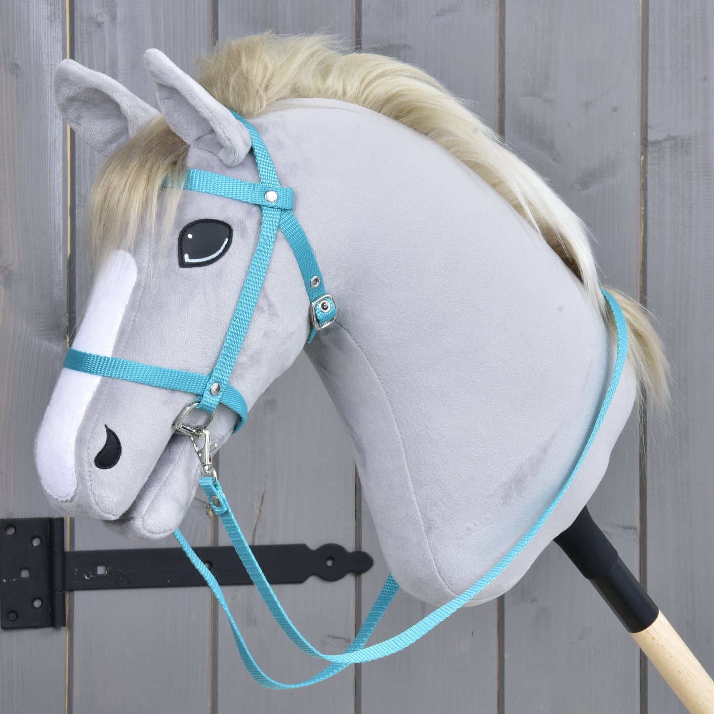 Ein graues plüschiges Hobby Horse mit heller Mähne, das das Hobby Horse LarDen Zaumzeug mit Zügel aus Gurtband Turquoise trägt, ist auf einem Holzstab montiert und vor einem grauen Holzzaun platziert.