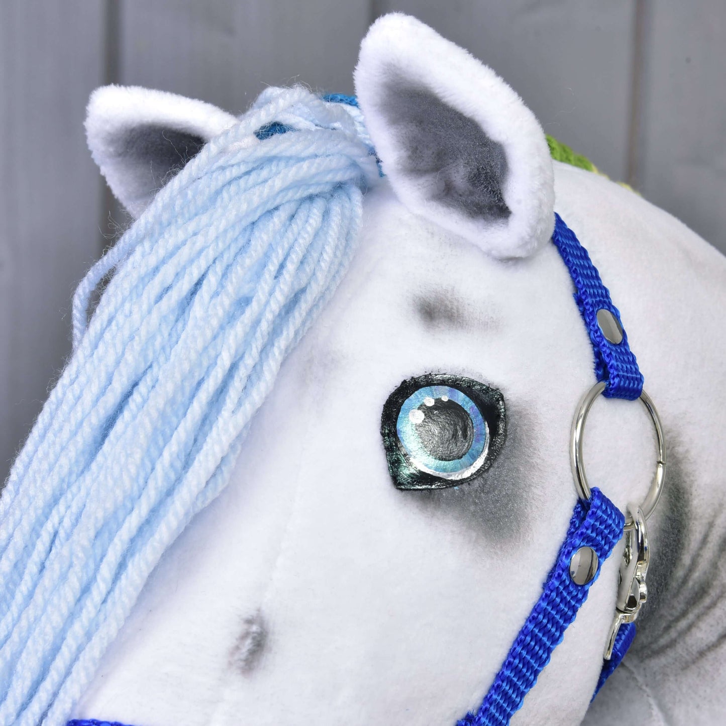 Nahaufnahme von Hobby Horse Rainbow mit Blauem Halfter von Hobby Horse LarDen mit weißem Plüschkörper, hellblauer Garnmähne, blauem Halfter und großem gesticktem Auge.