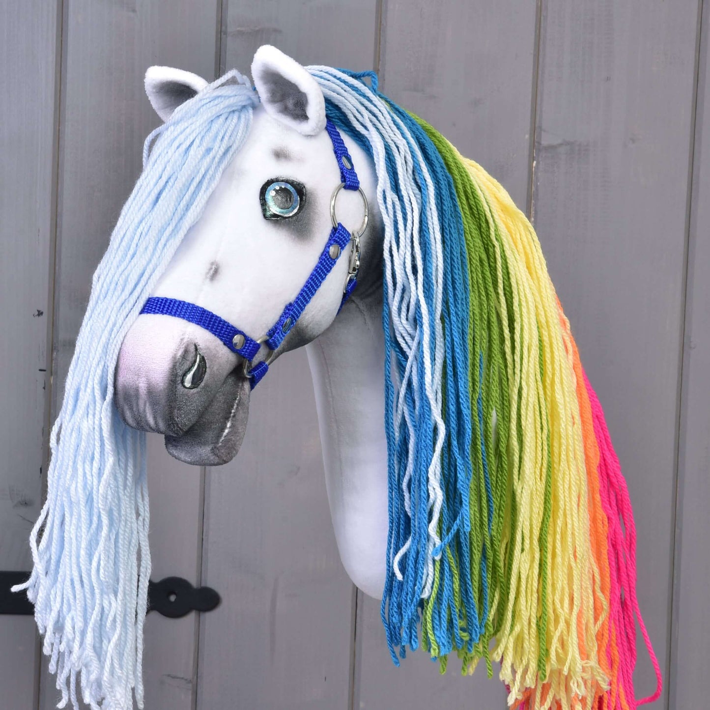 Hobby Horse LarDen Hobby Horse Einhorn Rainbow mit Trense und Halfter mit weißem Gesicht, blauer Trense und blauen Augen sowie langer pastellfarbener Regenbogenmähne in Blau, Grün, Gelb und Rosa vor grauem Holzhintergrund.