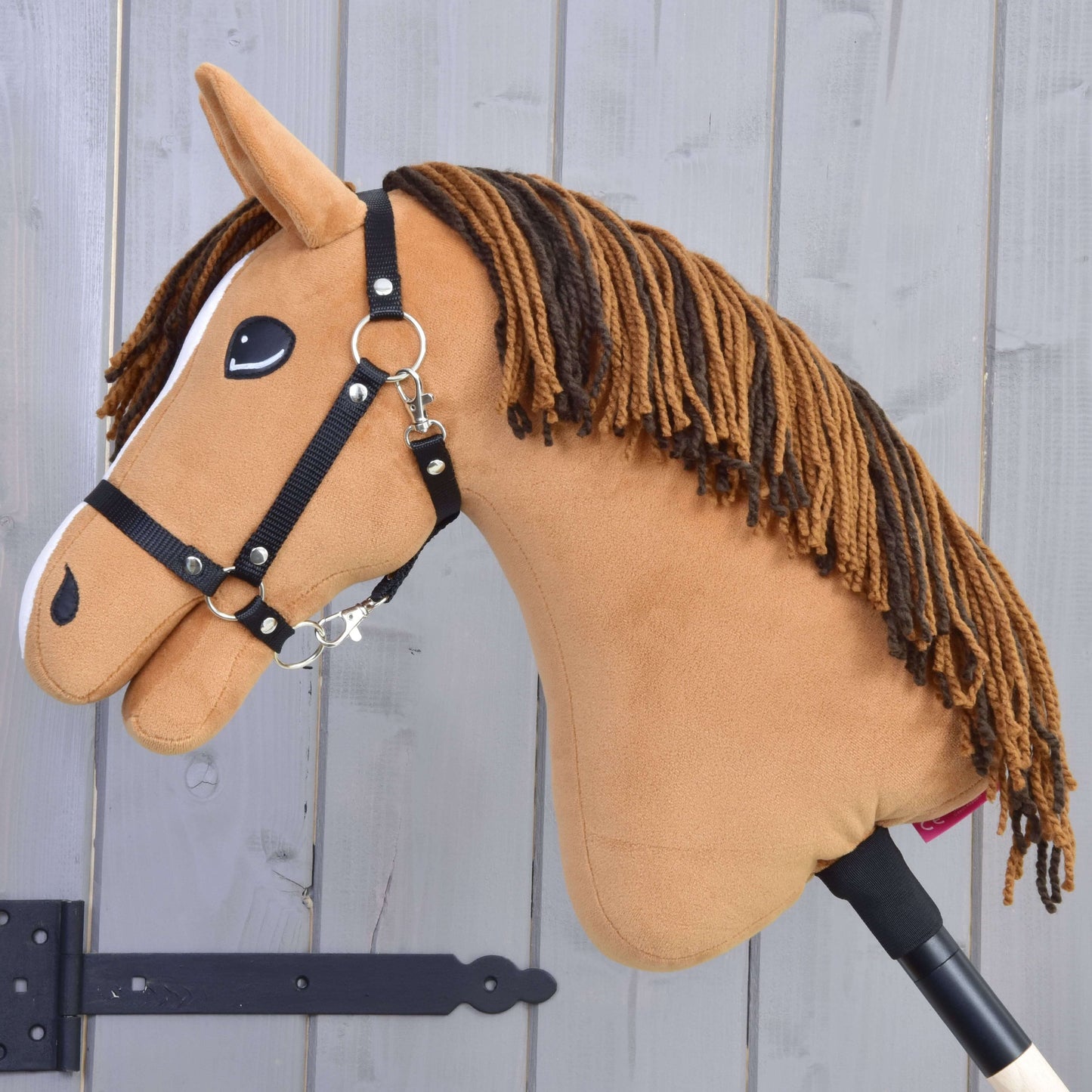 Hobby Horse Edward-LD mit Schwarzem Halfter von Hobby Horse LarDen mit plüschigem fuchsfarbenem Körper, brauner Garnmähne, schwarzem Halfter und weißen Akzenten steht vor einem hellgrauen Holzzaun.