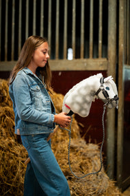 Lifestyle photo: a girl in a denim jacket stands in front of straw bales in a stable holding the Hobby Horse Elektra with Black Halter by Hobby Horse LarDen - white dappled plush head, blue eye, white yarn mane - shown here with the Graphite Full fur halter in leatherette and the knitted lead rope.