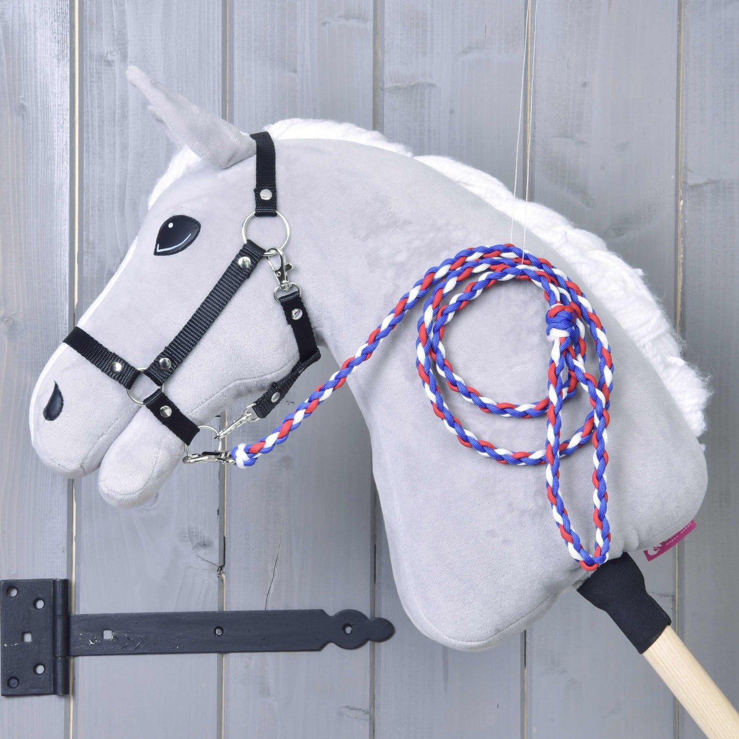 Hobby Horse LarDen Hobby Horse - rope stripe Flag CZ zeigt einen grauen Körper, weiße Mähne, schwarzes Zaumzeug und eine geflochtene Leine in Rot, Weiß und Blau auf einem Holzstab, gezeigt angelehnt an eine hellgraue Holzwand.
