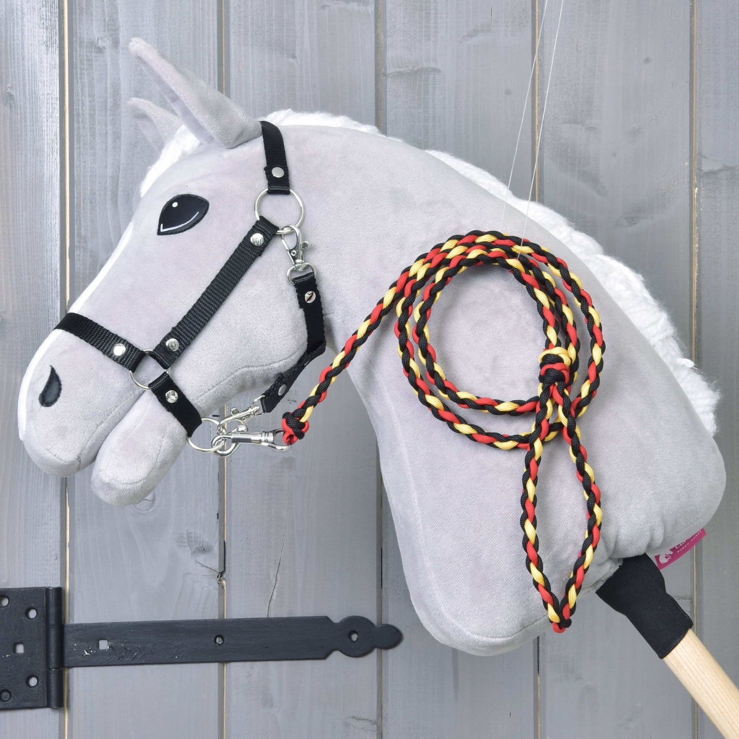 Hobby Horse LarDen „Hobby Horse - Führstrick Flag“ ist ein graues Plüsch-Hobby Horse mit Stabkörper, weißer Mähne, schwarzem Zaumzeug und einer geflochtenen Leine in Rot, Gelb und Schwarz, gezeigt vor einem Holz-Hintergrund.