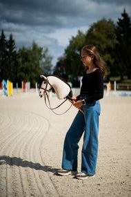 Mädchen im schwarzen Oberteil steht auf einem Sandplatz unter stürmischem Himmel und hält das Hobby Horse Frederik LD mit schwarzem Halfter von Hobby Horse LarDen, dahinter farbige Sprungstangen – cremefarbener Plüschkopf, dunkelbraune Garnmähne – hier mit dem vollständigen braunen Zaumzeug mit Reithalfter
