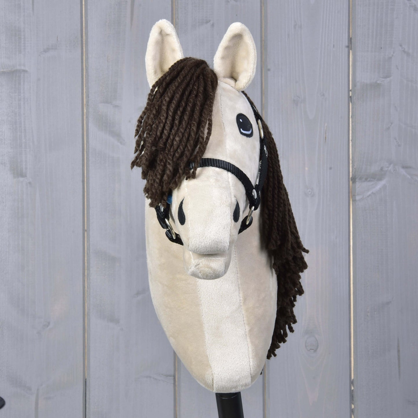 Hobby Horse Frederik LD mit Schwarzem Halfter von Hobby Horse LarDen steht aufrecht vor einem hellgrauen Holzzaun und hat einen beigen Körper sowie eine braune Garnmähne.