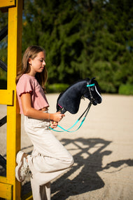 Lifestyle photo: a girl in a pink top leans against a yellow show jump wing holding the Hobby Horse Freya with Black Halter by Hobby Horse LarDen - black plush head, white blaze, black yarn mane - shown here with the complete turquoise bridle without noseband.
