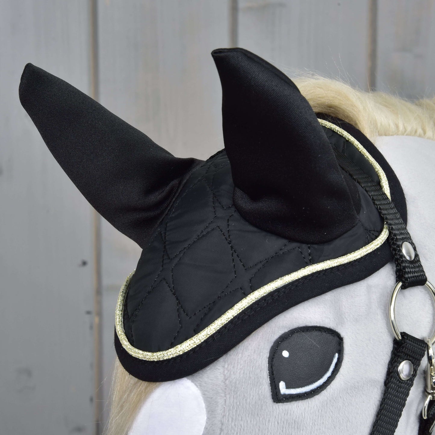 Gros plan sur la tête d’un cheval-jouet gris portant le bonnet d’oreilles noir matelassé de l’ensemble Majestueux de Hobby Horse LarDen avec bordure dorée, avec oreilles dressées et bride noire simple, présenté sur un fond en bois.