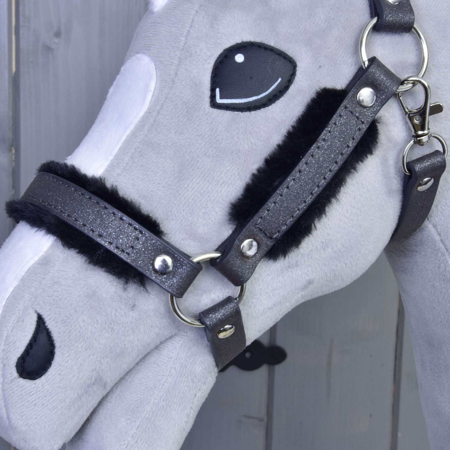 Gros plan sur la tête d’un Hobby Horse en peluche gris et blanc, présentant le licol avec fourrure - similicuir Graphite complet de Hobby Horse LarDen et des détails du visage cousus, sur fond en bois.