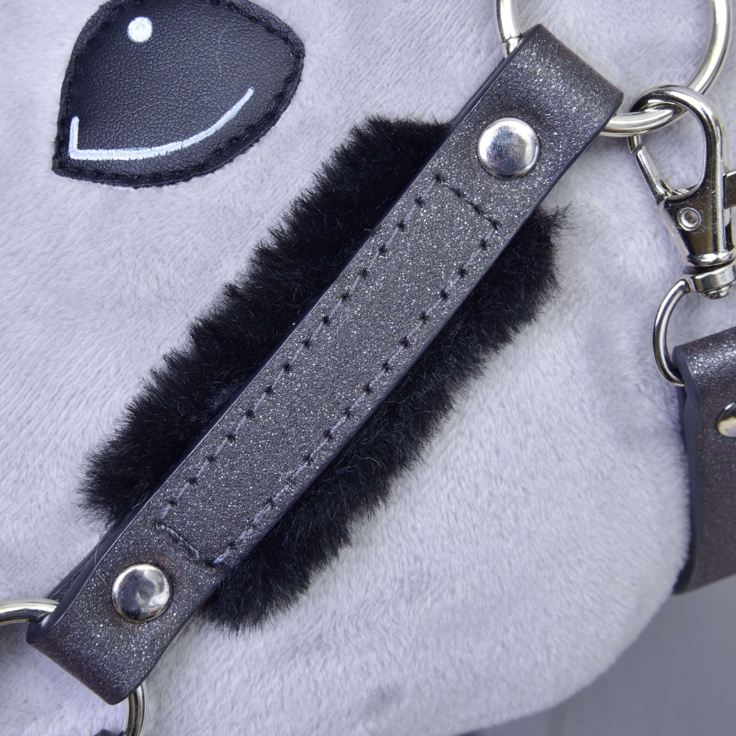 Gros plan sur le licol avec fourrure - similicuir Graphite complet de Hobby Horse LarDen, montrant un tissu en peluche gris, une bande noire duveteuse, des éléments métalliques, des finitions en similicuir et une partie d’un œil brodé souriant.