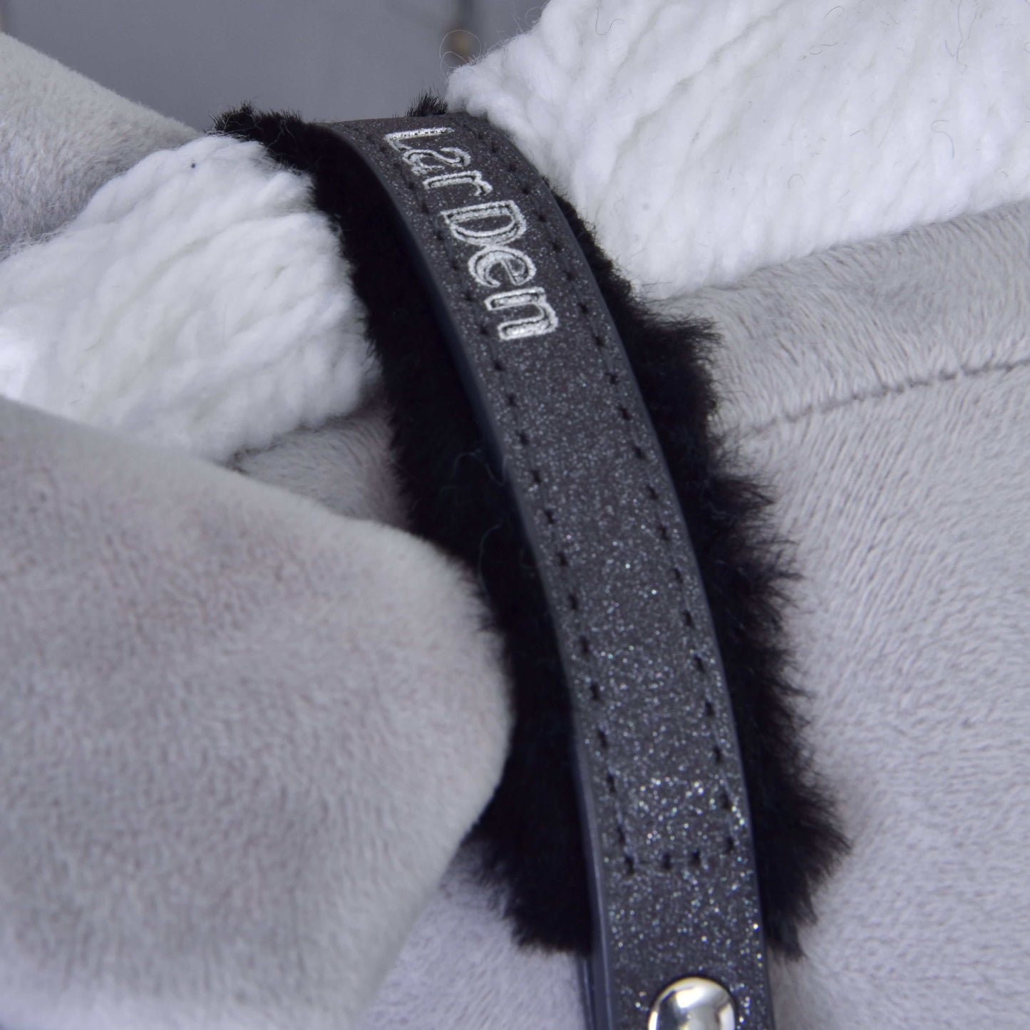 Gros plan sur le licol avec fourrure - similicuir Graphite complet de Hobby Horse LarDen, avec une sangle noire pailletée à broderie blanche, enroulée autour d’une corde en peluche blanche et posée sur un tissu doux gris clair.