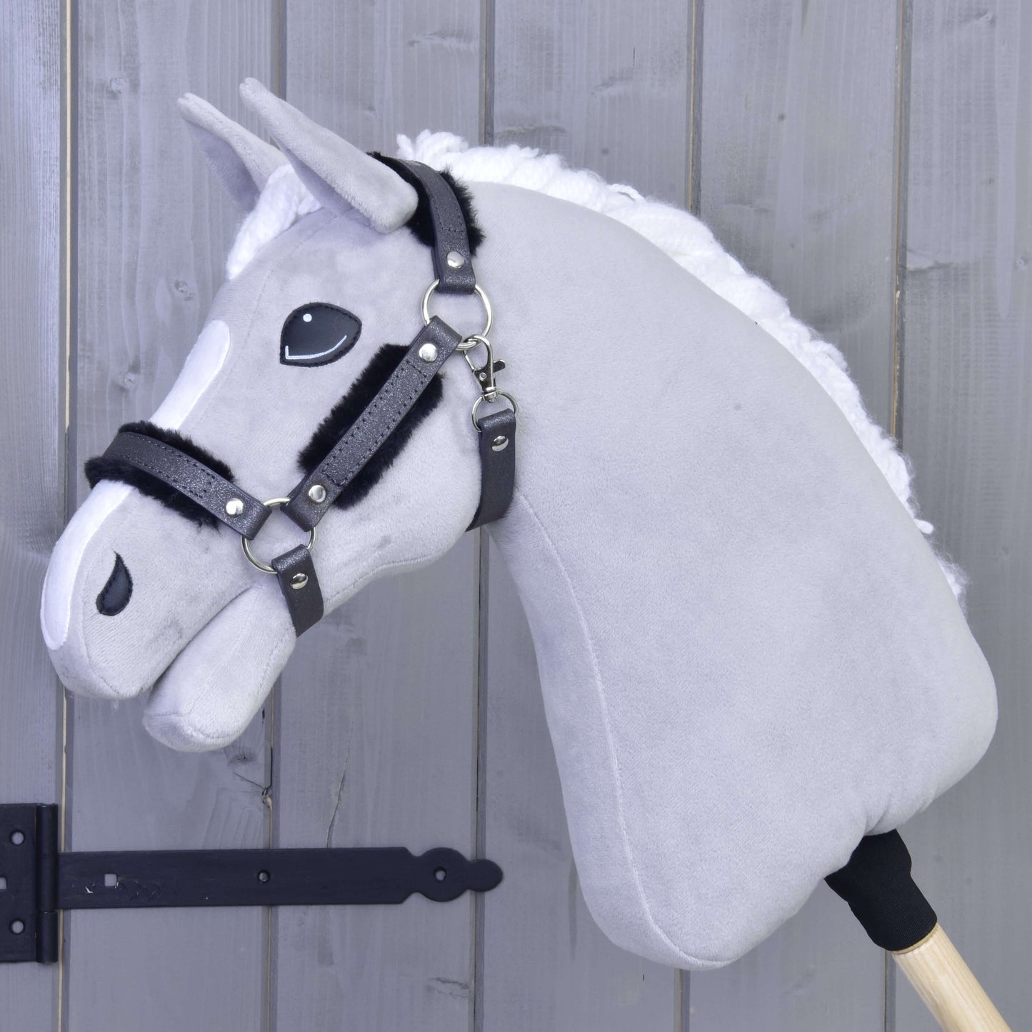 Licol avec fourrure - similicuir Graphite complet de Hobby Horse LarDen, monté sur un bâton en bois et appuyé contre un mur en bois gris clair, avec tête en peluche grise, crinière blanche, bride noire et licol en similicuir.