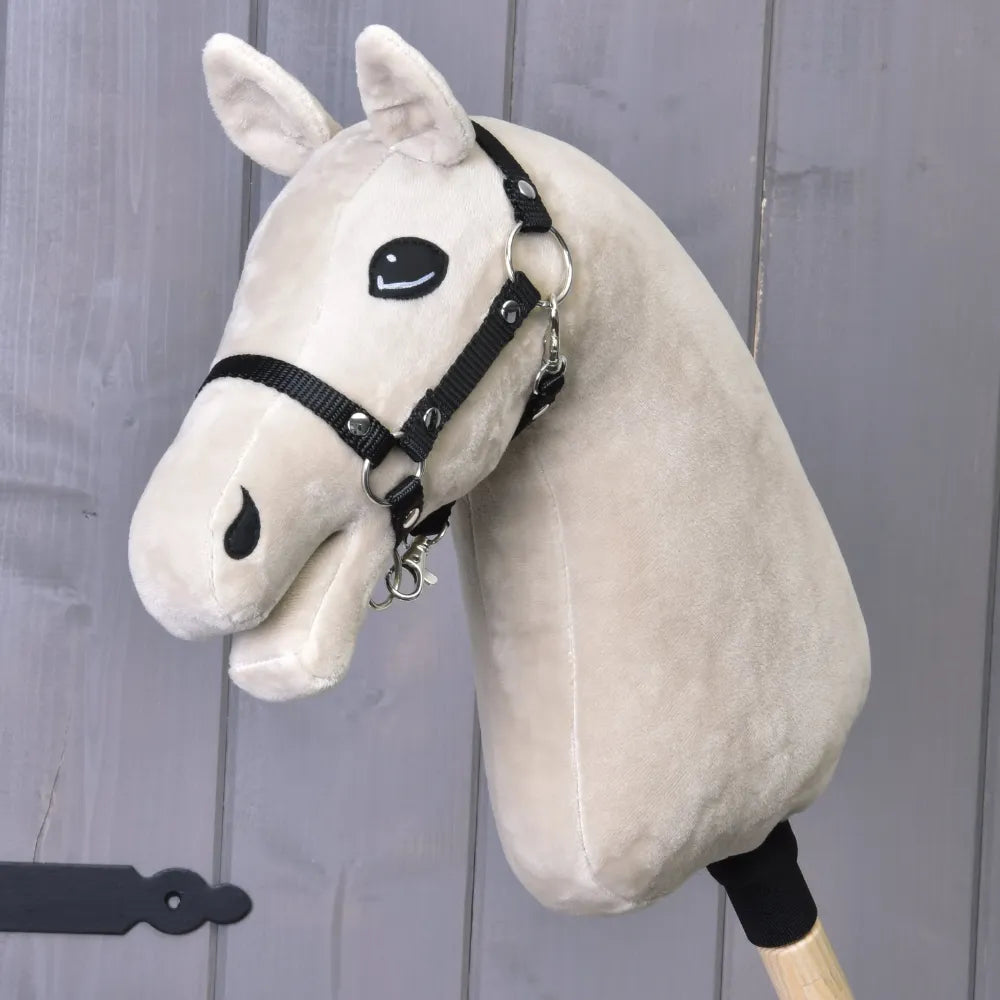 Construis ton cheval, tête de Hobby Horse en peluche beige clair avec crinière blanche, bride noire et bâton en bois, présentée sur un fond en bois gris.