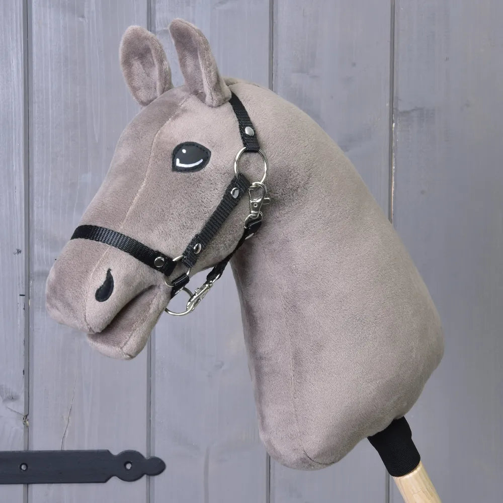 Baue dein Pferd Hobby Horse mit plüschigem taupefarbenem Kopf, blauen Augen und schwarzem Zaumzeug, auf einem Holzstab vor einer grauen Holzwand.