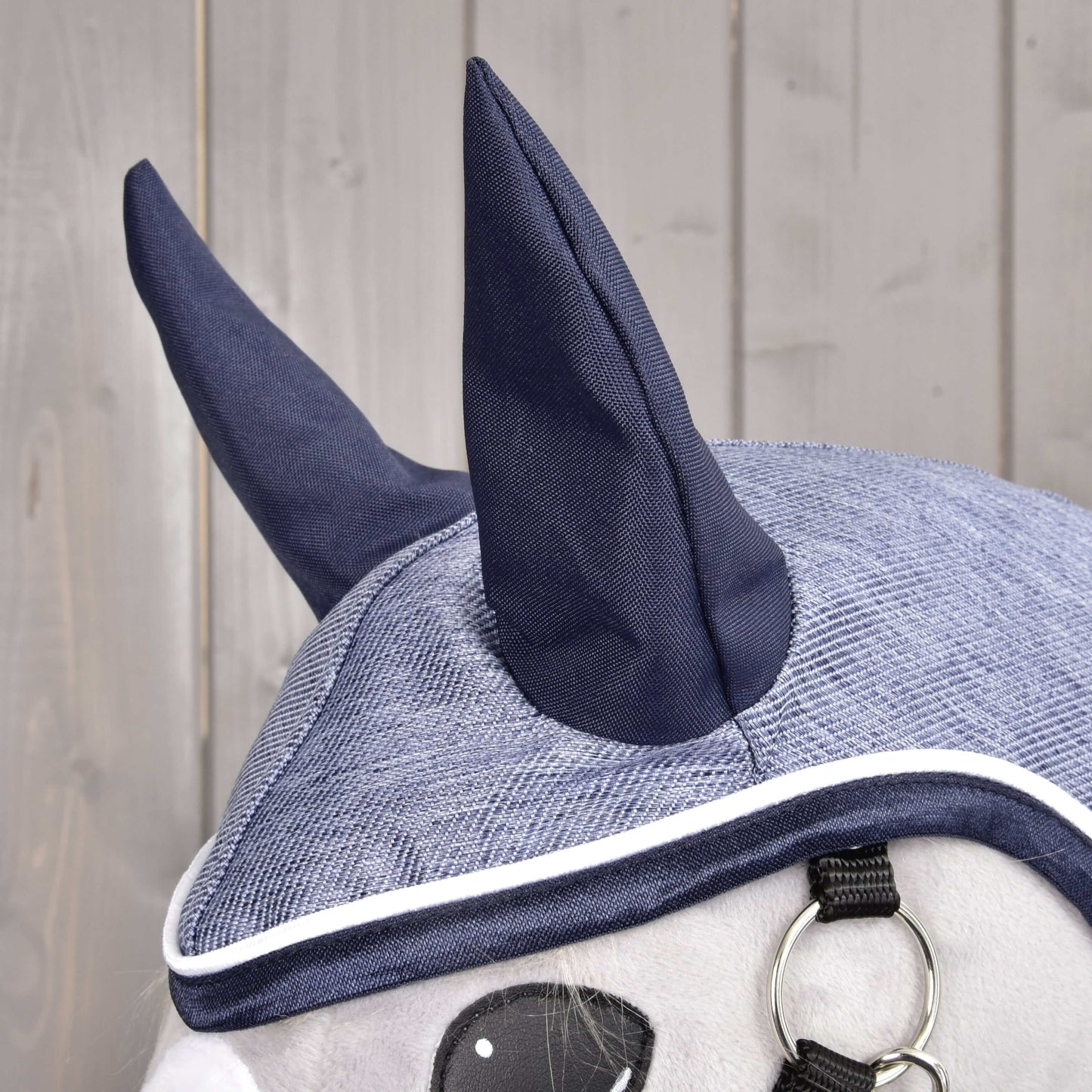 Nahaufnahme der grauen und dunkelblauen Mütze Regendecke Jeans von Hobby Horse LarDen mit zwei spitzen Ohren, weißer Einfassung und einem Metallring – ideal zum Kombinieren mit der Allwetter-Regendecke deines Hobby Horse – vor Holz-Hintergrund.