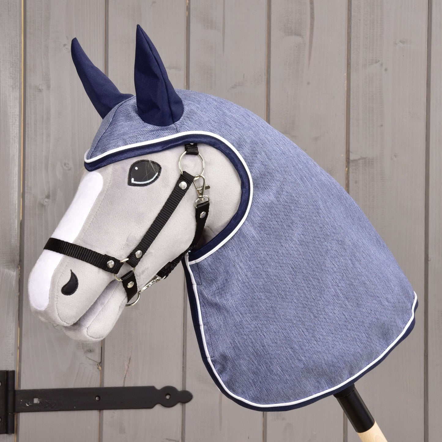 Regendecke Jeans von Hobby Horse LarDen an einem Plüschpferdekopf mit schwarzem Zaumzeug und blauer Regendecken-Kapuze mit Ohrenabdeckungen vor Holz – ideal als fantasievolles Spiel bei jedem Wetter.