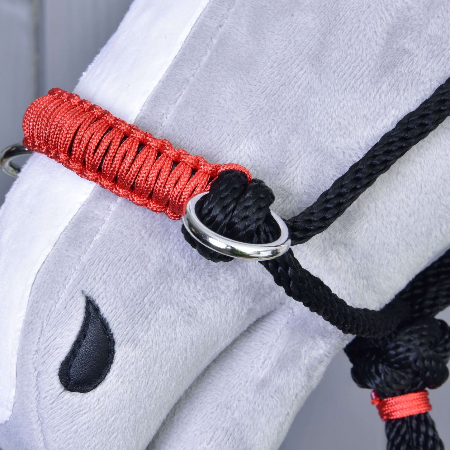 Gros plan sur la tête d’un cheval en peluche gris portant le Licou en corde Spark de Hobby Horse LarDen en rouge et noir avec un anneau métallique, ainsi qu’une broderie noire en forme de goutte près de l’œil. Idéal comme accessoire unique pour Hobby Horse.