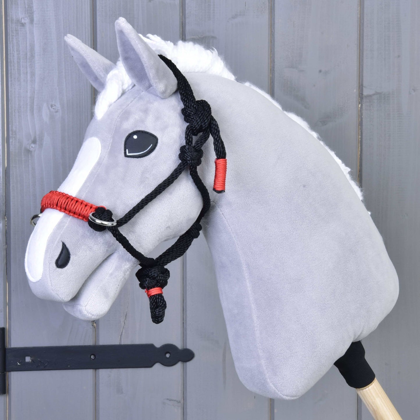 Licou en corde Spark de Hobby Horse LarDen sur un cheval jouet Hobby Horse gris avec crinière blanche et licou en corde noir et rouge, monté sur un bâton en bois devant un mur en bois gris.